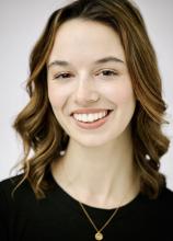 A young woman smiling, wearing a black t-shirt and gold necklace.