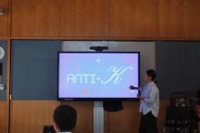 A woman is presenting in front of a large screen that displays the text "ANTI-K".