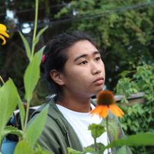 A person wearing a white t-shirt and olive green hoodie looks off to the side. The background is an outdoor setting with trees and flowers in the foreground. 