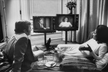 Black and white photo of a man and a woman sitting in front of two television sets. 