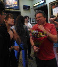 Man in red tshirt holding a bouquet of flowers speaks with several people.  