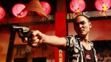 Man holding a gun in his right hand, standing in a room decorated with red lanterns. 