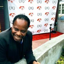 Charles Mudede leans over a concrete wall, with a red carpet and an Orcas Island Film Festival backdrop in the background.  
