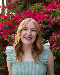 Head shot of woman with shoulder length light blonde hair wearing light green dress stands in front of a tree with deep pink flowers in the background.