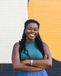 Photograph of Professor Golden M. Owens. She is a Black woman with long natural hair. She is looking at the camera and smiling. She is wearing a green top and blue pants. Her arms are folded, and there is a gold watch on her left wrist. 