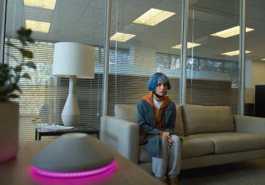 Screenshot from KIMI.  Female sits on a couch in an office, looking at a virtual assistant device on the table. 