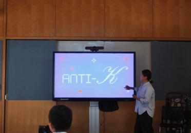 A woman is presenting in front of a large screen that displays the text "ANTI-K".