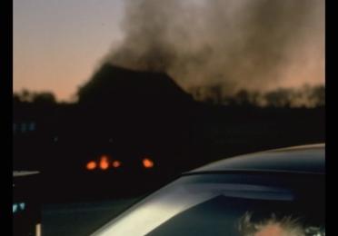 Filmmaker David Lynch lighting a cigarette while seated in a car. A large plume of dark smoke rises in the background. 