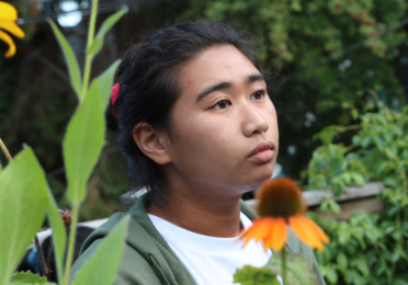 A person wearing a white t-shirt and olive green hoodie looks off to the side. The background is an outdoor setting with trees and flowers in the foreground. 