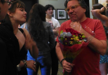 Man in red tshirt holding a bouquet of flowers speaks with several people.  