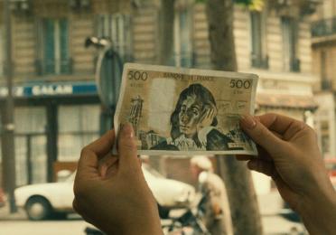 A person is holding a 500 French Franc banknote. The background appears to be a street scene in a city.