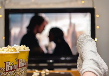 Person watching a movie at home, with a popcorn container on the table and stockinged feet resting on the edge of the table.