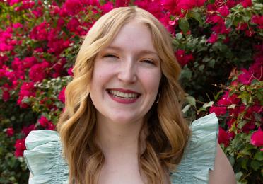 Head shot of woman with shoulder length light blonde hair wearing light green dress stands in front of a tree with deep pink flowers in the background.