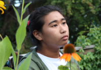 A person wearing a white t-shirt and olive green hoodie looks off to the side. The background is an outdoor setting with trees and flowers in the foreground. 