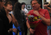 Man in red tshirt holding a bouquet of flowers speaks with several people.  