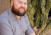 Man with beard wearing a grey shirt sits looking at the camera.