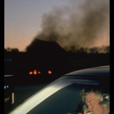 Filmmaker David Lynch lighting a cigarette while seated in a car. A large plume of dark smoke rises in the background.
