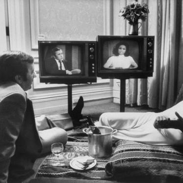 Black and white photo of a man and a woman sitting in front of two television sets.